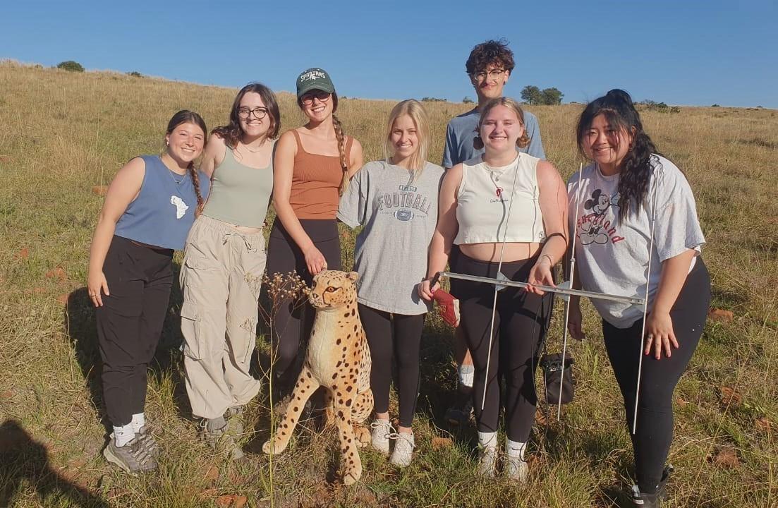 A group of smiling students poses with a cheetah stuffed animal, facing the camera in a cheerful setting